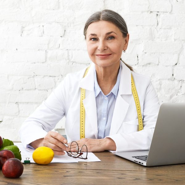 Indoor shot of positive smiling Caucasian female nutritionist in her sixties working online, sitting in front of open laptop pc, consulting clients on nutrition via social networks, making notes
